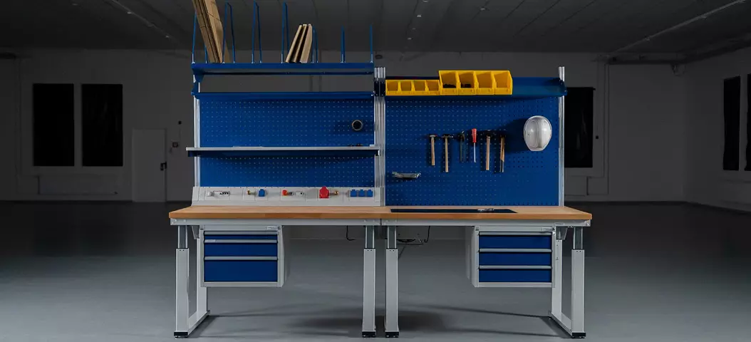 Bild einer elektrisch höhenverstellbaren Werkbank mit blauen Lochplatten zur Werkzeugorganisation und mehreren Schubladen, in einer großen, leeren Werkstatt oder Industrieumgebung.