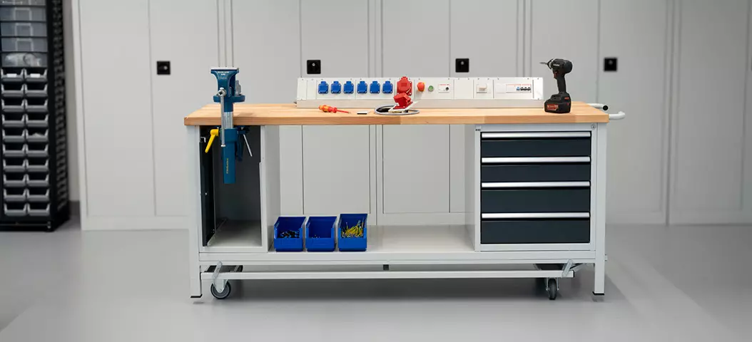 Absenkbare Werkbank mit Holzarbeitsplatte, ausgestattet mit Schubladen, Werkzeugen und blauen Lagerkästen in einer sauberen Werkstattumgebung.