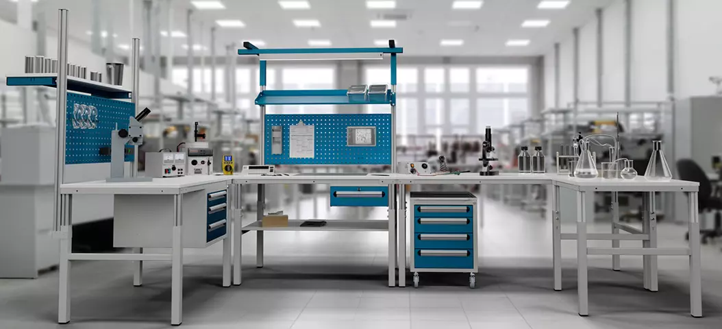 Umfassende Laborarbeitsstation Serie E in Eckkonfiguration, ausgestattet mit Tischen, blauen Schubladen und einer Rollwerkbank, sowie einem Systemaufbau für technische Instrumente in einem hellen Industrielabor.