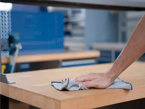 Hand reinigt eine robuste Holzarbeitsplatte mit einem Tuch in einer professionellen Werkstattumgebung.