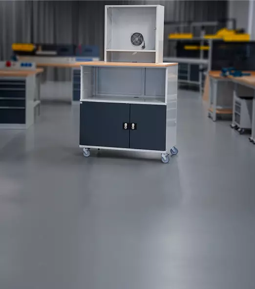 Mobiler Computertisch auf Rollen mit mehreren Ablagen und grauem Monitorschrank in industrieller Umgebung.