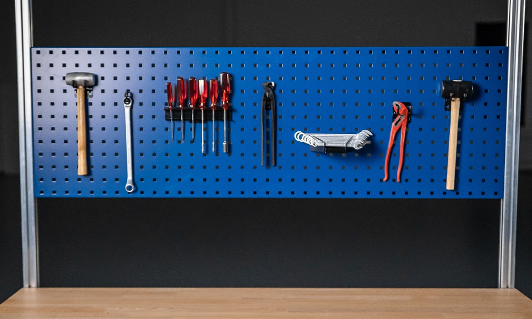 Werkzeugwand mit blauer Lochplatte, auf der verschiedene Werkzeuge wie Hammer, Schraubendreher, Schraubenschlüssel, Zange und Inbusschlüssel ordentlich aufgehängt sind, vor einer Holzarbeitsfläche.