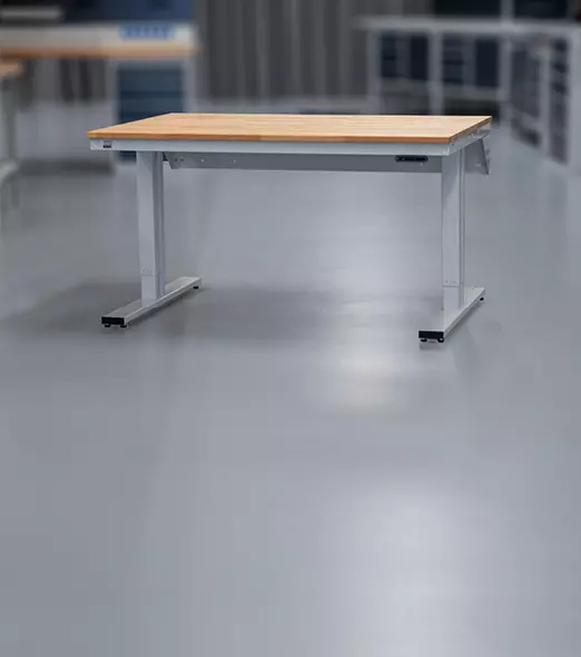 Elektrisch höhenverstellbarer Werktisch mit heller Holzplatte undhellgrauem Gestell auf Rollen in heller Werkstatt industrieller Umgebung.