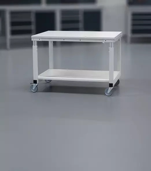 Mobiler Tischwagen in hellgrau mit einer unteren Ablagefläche und Rollen in einer industriellen Umgebung. 