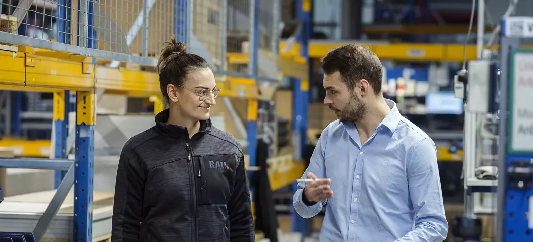 Zwei Mitarbeiter in einem Lagerhaus, die lächelnd miteinander sprechen, Mann in Hemd zeigt auf etwas außerhalb des Bildes, während Frau in Arbeitskleidung aufmerksam zuhört.