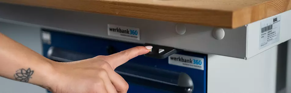 Eine Person betätigt den Knopf eines Bedienelements einer Werkbank von werkbank360, wobei der Fokus auf der Handaktion und der Markenkennzeichnung liegt.
