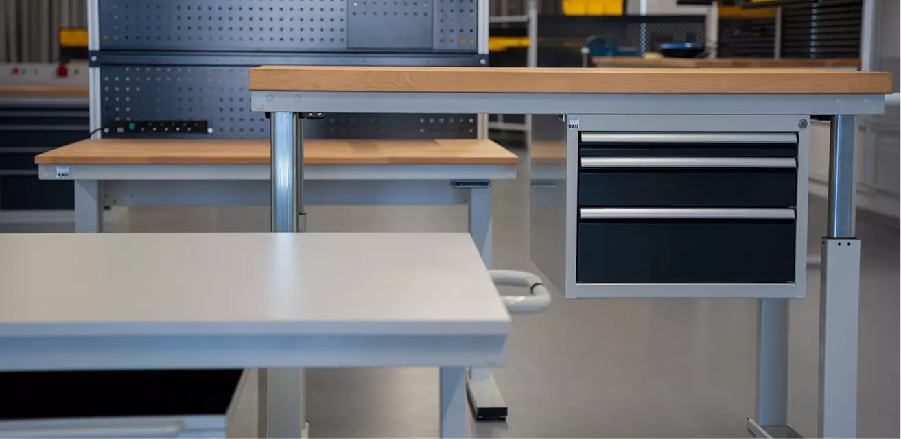 Reihe elektrisch höhenverstellbarer Arbeitstische in unterschiedlichen Ausführungen mit Schubladen, in einer Werkstattumgebung.