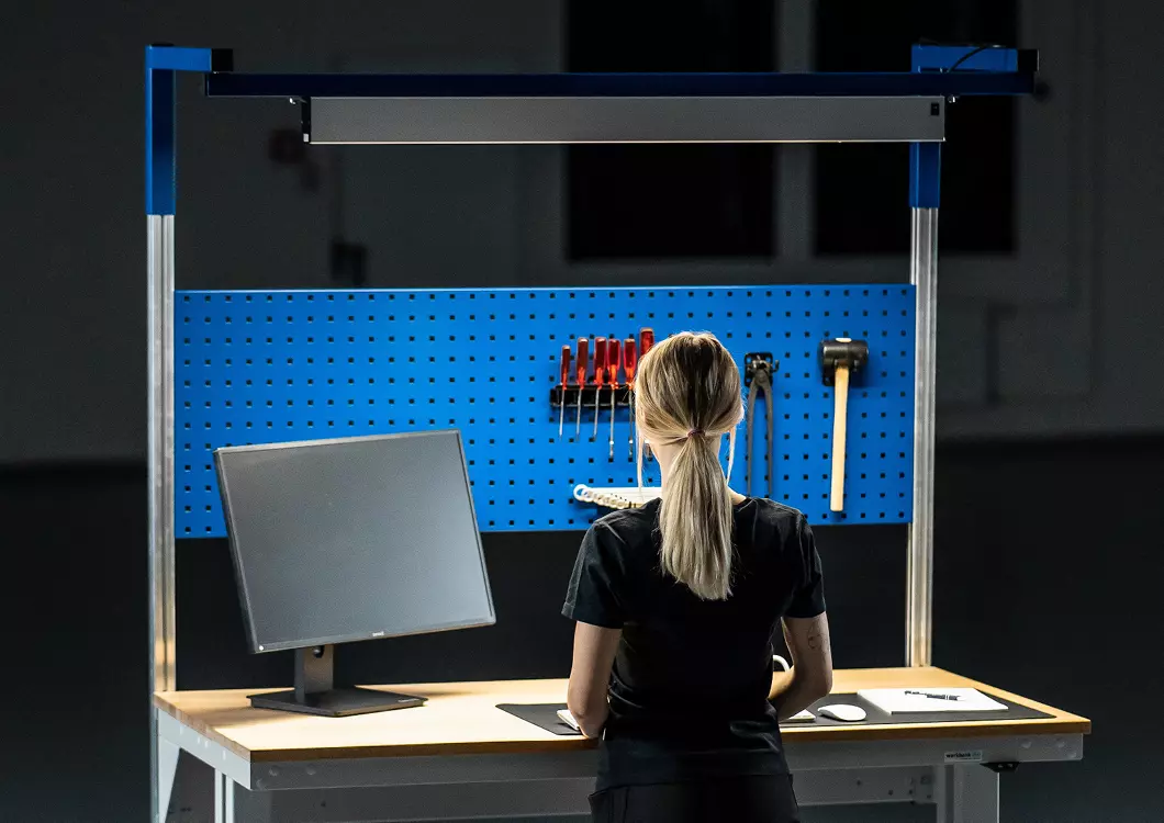 Frau steht an einem Arbeitsplatz mit montiertem Systemaufbau, mit einer blauen Lochplatte mit Werkzeug und einer LED-Lampe.
