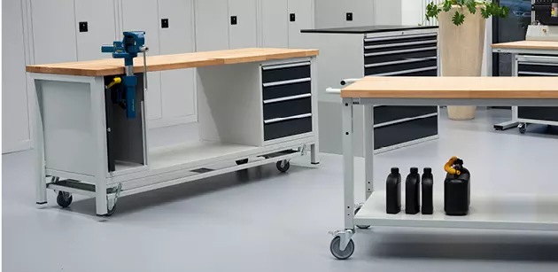 Mobil einsetzbare Werkbänke mit Holzarbeitsplatte und grauem Metallgestell auf Rollen, ausgestattet mit Schraubstöcken und Schubladen, in einer Arbeitsumgebung.