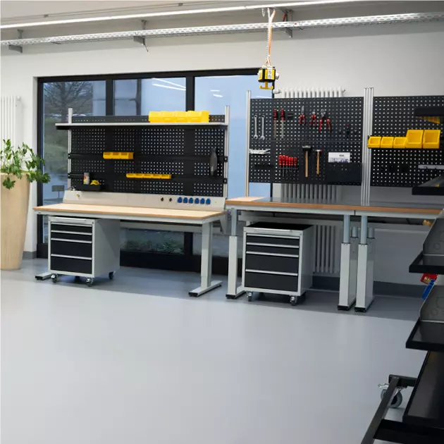 Moderne Werkstatteinrichtung mit zwei Werkbänken, ausgestattet mit Lochwänden, Werkzeughaltern und gelben Sichtlagerkästen, in einem hellen Raum mit Fensterblick.